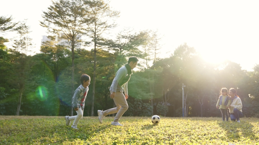 Asian father and son playing soccer for fun outdoors in park while mother and daughter watching from behind