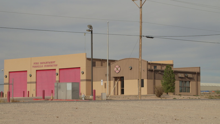 Establishing shot of a fire station in a dusty, rural setting of Tornillo, Texas, a border-town in Texas near Mexico.  Three red doors on a brown building on a sunny day with dusty roads.