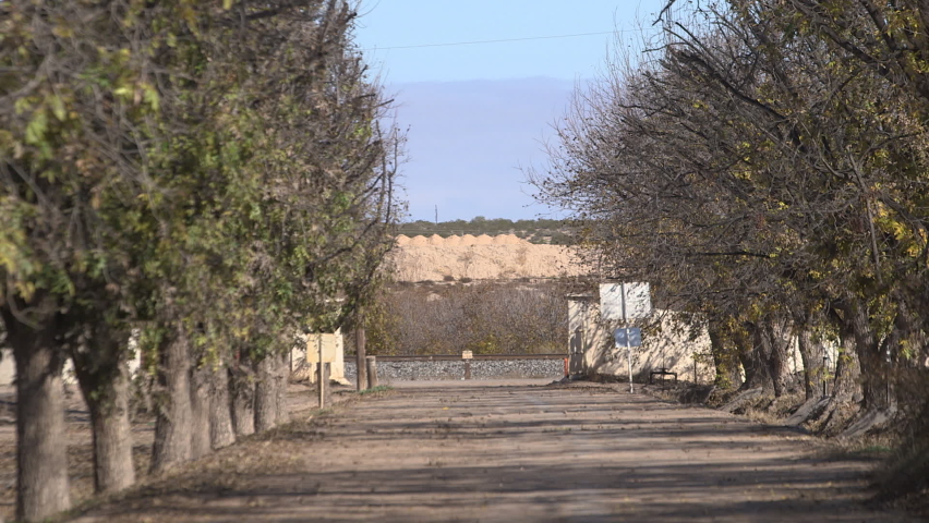 A long driveway at a pecan tree ranch in Tornillo, Texas as cars pass on a small highway.  Dusty grounds, trees, and mountains are in the background of this rural border-town near Mexico.