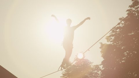 SLOW MOTION: Young man walking on slackline between the houses in big urban city at golden sunset