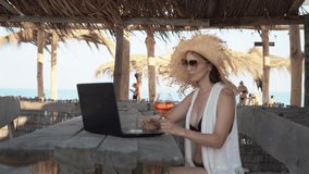 a beautiful Caucasian woman in a straw hat sits at a table in a tropical bar drinking a cocktail and makes a video call using her laptop and an app.