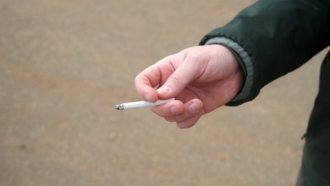 Tobacco smoke addiction, unhealthy lifestyle concept. Man's hand with a smoldering cigarette smoking a bad habit. Man is holding a burning cigarette while smoking.
