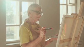Senior Artist, Art Workshop, Sun Rays, Painting Woman. Happy senior woman artist paints a picture on canvas while standing in a sunny workshop - Powered by Shutterstock - Get 15% off with code: PIKWIZARD15