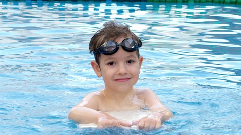 Cheerful kid in goggles bathing in the pool. A preschool child in the pool in swimming goggles splashes water in the pool, a boy jumps and holds a swimming board in his hands.