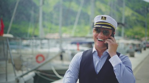 Happy smiling ship captain walking in marina and speaking to cell phone. Summer vacation at sea, sailing and travelling concept.