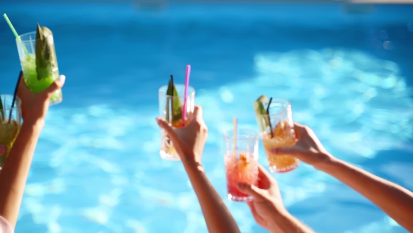 Back view of friends clinking glasses with cocktails sitting by swimming pool on sunny summer day. People have fun at poolside party toasting, drinking beverages at luxury tropical villa. Slow motion. | Shutterstock HD Video #1076893742