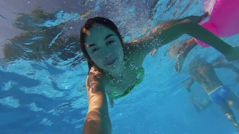 Fitted pretty woman in swimsuit diving under water in swimming pool, shooting herself on action camera. Mixed race girl filming selfie video bathing underwater. Partying with friends on beach party.