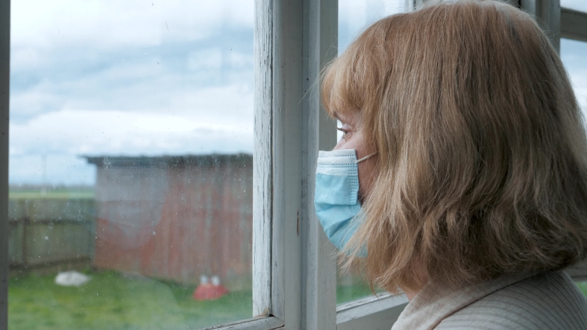Mature Woman In Medical Mask On Looks Out At Street With Her Hand On Glass