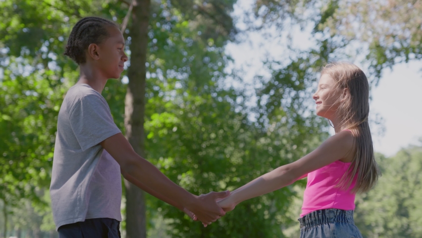 Side view of caucasian girl and african boy hold hands standing together in park. Cute diverse teenage friends holding hands and looking at each other outdoors