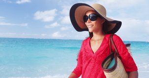 Luxury beach vacation elegant lady walking on beach stroll with beachwear sun hat and straw tote bag wearing red cover-up dress. SLOW MOTION VIDEO