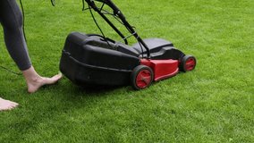 Caucasian woman mowing green grass lawn with electric grasscutter mower sunny summer day, high angle view, rear side view