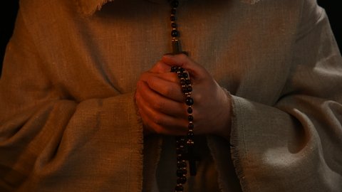 Close up senior priest or monk with gray bristle beard in haircloth cowl robe praying in candlelight, telling beads of finger rosary, front view