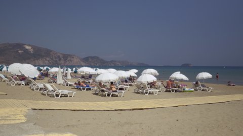 Zakynthos, Greece July 9 2021: Mediterranean terrain organized sandy beach with bathers. Day sunny view of Laganas seafront with calm sea and sea beds with white umbrellas under blue sky.