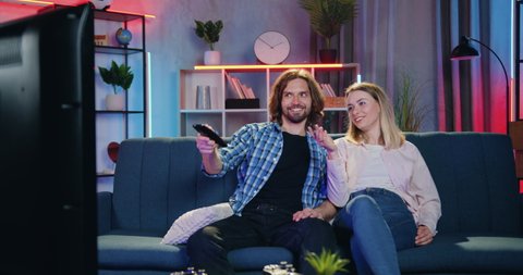 Adorable happy smiling loving couple sitting on the couch in living-room in front of TV and watching interesting program when their two kids running up to them with huggs
