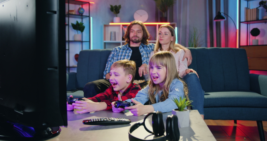 Attractive happy relaxed couple eating popcorn and talking between themselves while their son and daughter celebrating victory in videogames with raised hands,evening family leisure concept