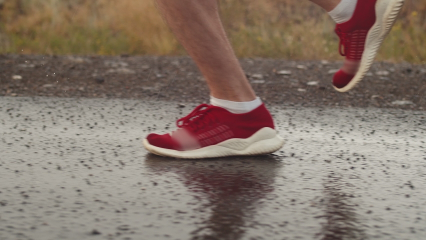 Man triathlete legs in red sneakers run on asphalt during rain, side view, closeup. Athlete runs along road during thunderstorm