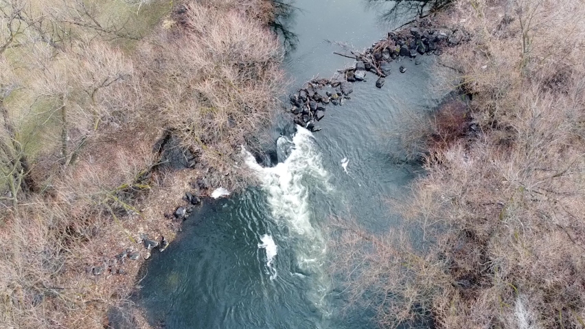 Small river dam and whitewater rapids flowing on scenic, idyllic, willow tree lined river in winter in rural countryside of Boise, Idaho, USA. Aerial drone static closeup