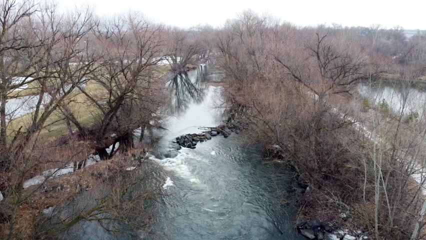 Small river dam and rapids on scenic, idyllic, willow tree lined flowing river in winter in rural countryside in Boise, Idaho, USA. Aerial drone reverse down river