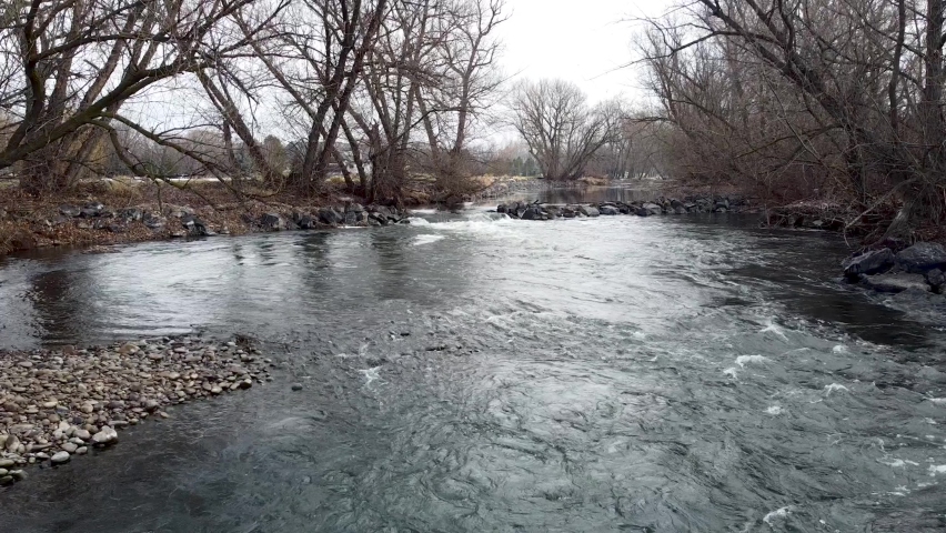 Small river dam and rapids on scenic, idyllic, willow tree lined river in winter in rural countryside in Boise, Idaho, USA. Aerial drone low closeup hovering above river