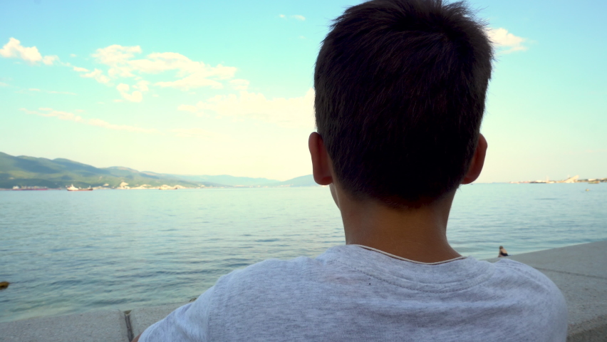 The boy stands on the embankment and looks at the sea. Tanned skin. Age 7-8 years old. Dreamer. Gray T-shirt. Blue sky.