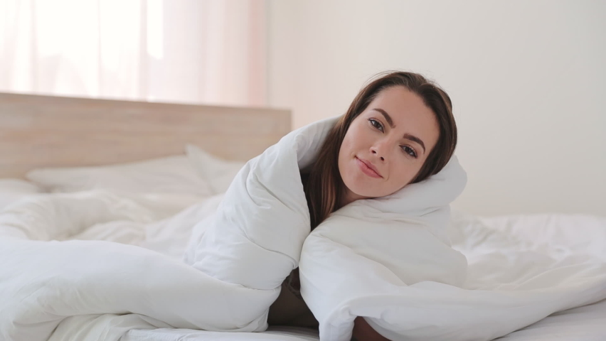 Happy brunette young woman in casual beige tank-top pajamas lying in bed wrapped in blanket look camera relax spend time in bedroom lounge home in own room house. Bedtime good mood lifestyle concept Royalty-Free Stock Footage #1076930717
