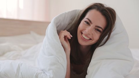 Happy brunette young woman in casual beige tank-top pajamas lying in bed wrapped in blanket look camera relax spend time in bedroom lounge home in own room house. Bedtime good mood lifestyle concept