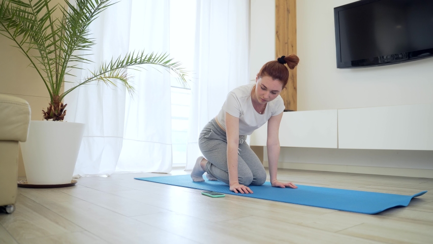 young redhead girl woman in casual clothes doing physical exercise using mobile phone app with workout. female on mat stretching Online tutorial of yoga, aerobics, fitness pilates at home living room