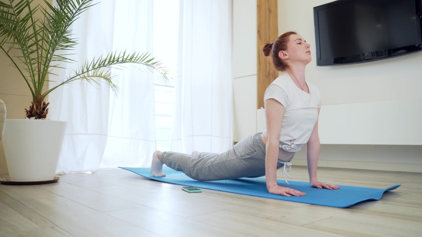 young redhead girl woman in casual clothes doing physical exercise using mobile phone app with workout. female on mat stretching Online tutorial of yoga, aerobics, fitness pilates at home living room
