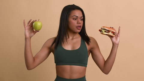 Healthy Food Choice. Sporty Black Lady Choosing Apple Instead Of Unhealthy Burger Posing Standing On Beige Background. Studio Shot. Dieting And Weight Loss Nutrition Concept. Slow Motion