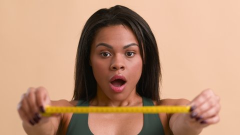 Weight Gain. Shocked Black Woman Showing Measuring Tape After Gaining Excess Kilograms Posing Over Beige Studio Background. Unsuccessful Slimming, Weight-Loss Failure Concept. Selective Focus