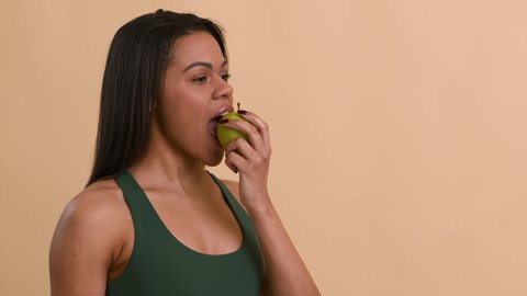Healthy Diet. Black Fitness Lady Eating Apple Posing Smiling To Camera Looking Aside Standing On Beige Studio Background. Side View Shot Of African American Woman Enjoying Apple Fruit. Weight Loss