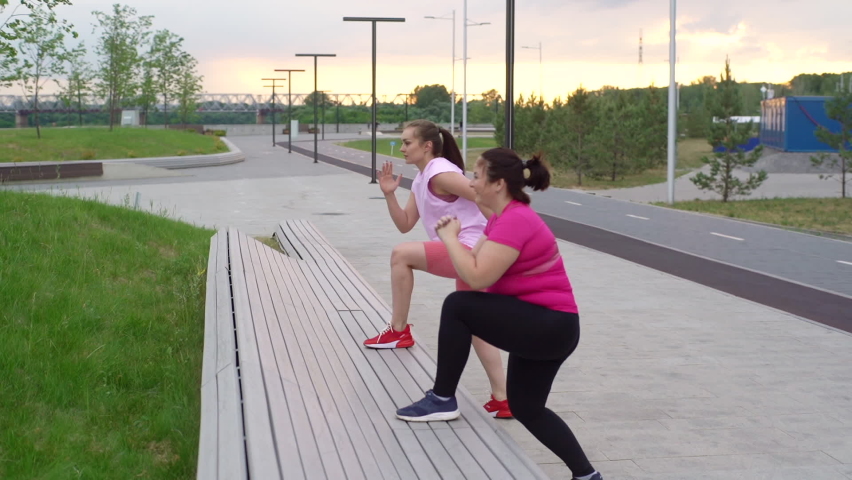 Overweight young woman doing single leg squats using bench with personal trainer outdoor in cloudy summer evening. Training from athletic lady for fat female with big abdomen wearing sportswear.