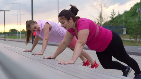 Personal training from positive athletic lady for obese female with big abdomen outdoor in cloudy summer evening. Fat caucasian woman performing push up exercise with female trainer using bench.