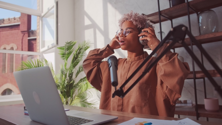 African American female wearing wireless headphones greets his followers and talking on microphone, working at communication webinar in living room. Black woman blogger social media putting on blog. Royalty-Free Stock Footage #1076941634