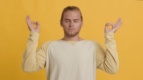 Harmony And Peace Concept. Handsome Young Man Meditating Over Yellow Background, Calm Millennial Guy Opening Eyes And Deeply Breathing, Keeping Hands In Mudra Gesture, Slow Motion Footage, Closeup