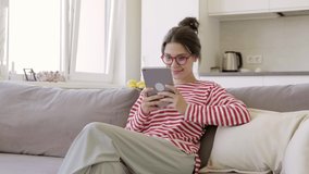 Modern young girl sitting on sofa hold tablet computer receive good news, gesturing with joy. Joyful female in glasses with pad device celebrating success, win, showing sincere emotions.