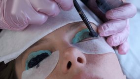 Cosmetologist puts black paint on woman's lashes using brush. Laminating eyelashes procedure in beauty salon. Beautician is painting lashes in black colour in cosmetology clinic, face closeup.