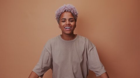 Young cheerful african american woman showing and pointing with fingers to healthy white teeth and broad smile has blonde hair wearing casual t-shirt over beige background. Dental health concept.