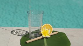 Video footage of glass with orange juice, bamboo straw, half of fresh orange, yellow frangipani flower and bubbling blue swimming pool on background.