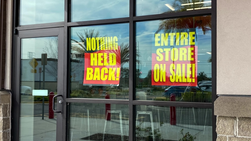 Zooming out on signs in the window of a retail store going out of business that say the entire store on sale, everything must go and nothing held back.