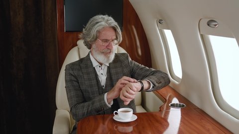 Busy mature business man looking on his watch and then at window while travels on private plane. Rich senior man drinking tea or coffee while enjoying luxury journey in first business class airlines