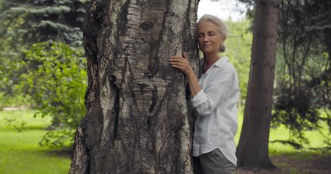 Side view of mature woman hugging tree and smiling outdoor. Portrait of elegant senior female embrace tree trunk in summer park