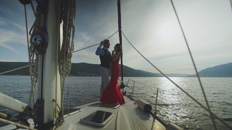 Romantic evening view of embracing couple standing on sailboat bow while cruising. Summer vacation at sea, sailing and travelling concept.