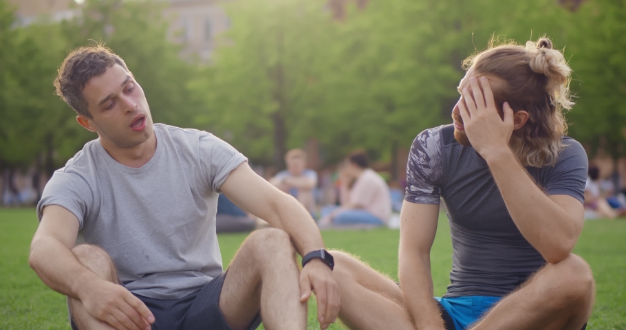 Young male friends relaxing on green lawn after exercise in park. Portrait of caucasian men resting on grass after group fitness training outdoors in city park