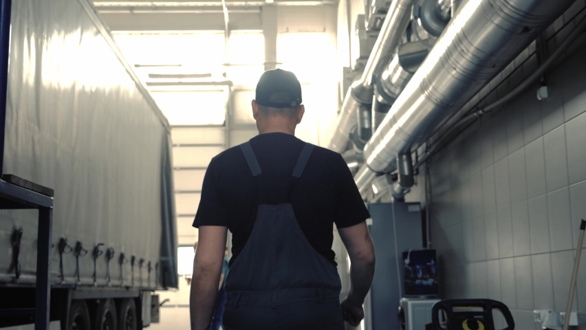 A mechanic in overalls walks through the workshop along a cargo trailer. Truck service station everyday life Royalty-Free Stock Footage #1076966240