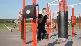 Street fighter in black clothes boxing in punching bag outdoors. Young man doing box training and practicing punches at outside gym. Male athlete exercising on sport ground. Sport motivation video