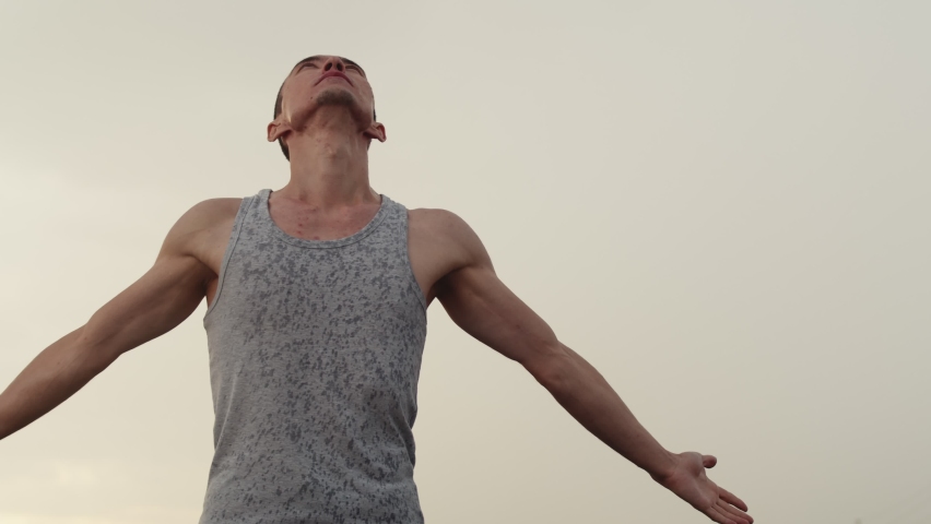 Man of athletic build in gray T-shirt is standing in outdoor against background of overcast sky, arms outstretched and looking up, enjoying rain, side and down view, slow motion