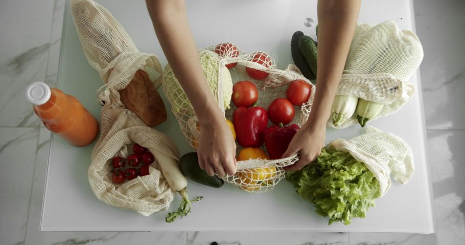 Woman takes yellow and red pepper out from reusable grocery bag with vegetables on a table at the kitchen at home after grocery shopping. Zero waste and plastic free concept. Mesh cotton shopper. Royalty-Free Stock Footage #1076979698