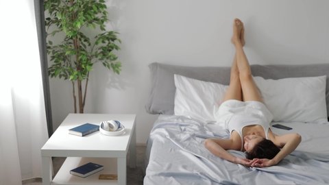 Relaxed woman in sleepwear lying in bed with legs on wall and enjoying sunny morning at bright bedroom. Laziness and people concept.