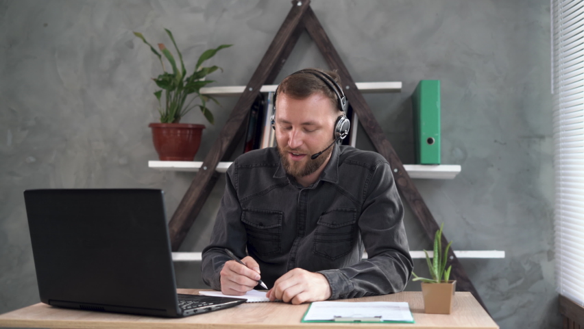 a remote worker, a bearded Caucasian man, sits at the workplace in the office dressed in headphones with a microphone, makes a video call and makes notes in a notebook. Royalty-Free Stock Footage #1076990372
